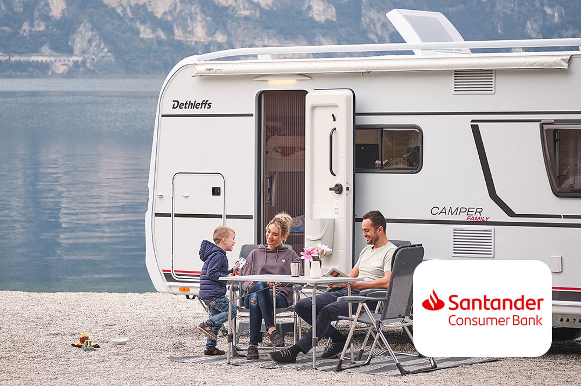 Genießen Sie maximale Flexibilität mit Ihrem Wohnwagen und Pkw. Eine Familie sitzt vor einem Wohnwagen vor einem See.