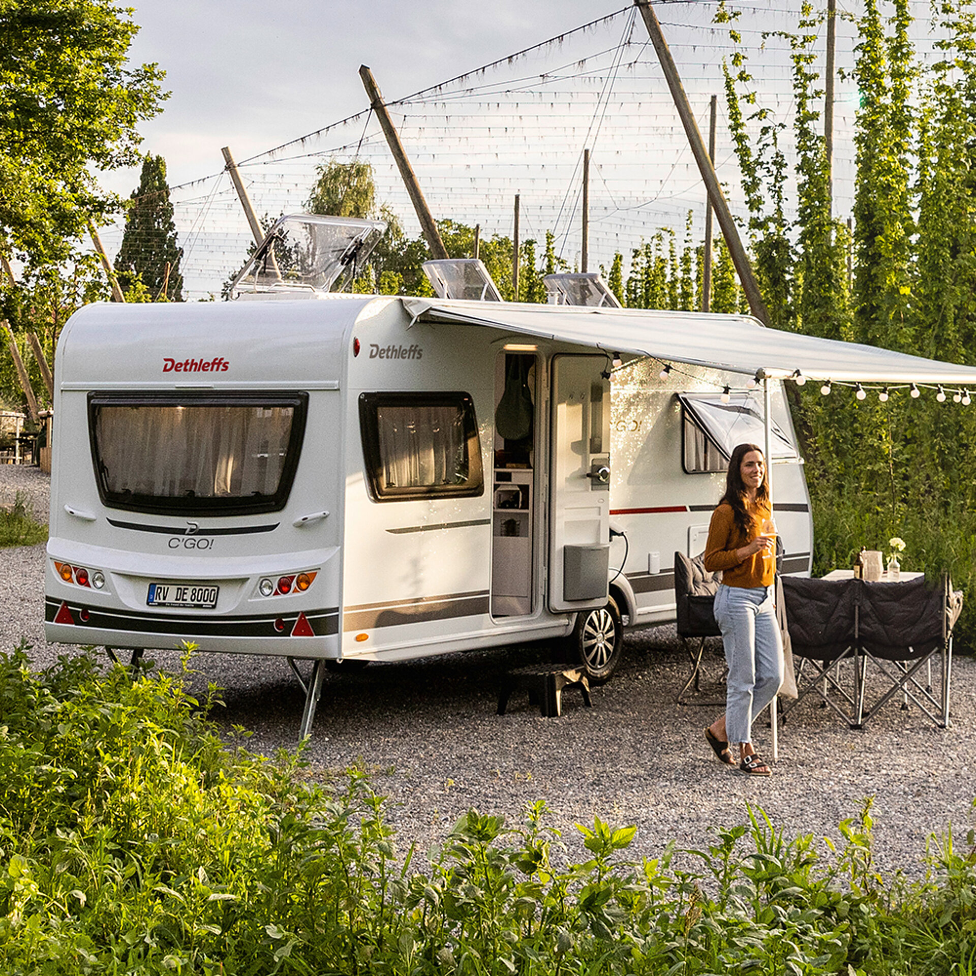 Wohnwagen in einem Gartenbereich mit aufgestellter Markise und zwei Menschen im Vordergrund.